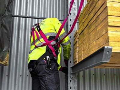 Person mit Warnweste und pinkem Auffanggurt arbeitet sicher neben Holzlager &ndash; zeigt Schutzma&szlig;nahmen im Industriebereich