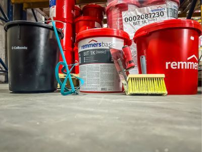 Rote Eimer mit &bdquo;Remmers BIT 1K&ldquo; und Reinigungswerkzeug stehen auf Boden, zeigen Bauchemie im Lager.